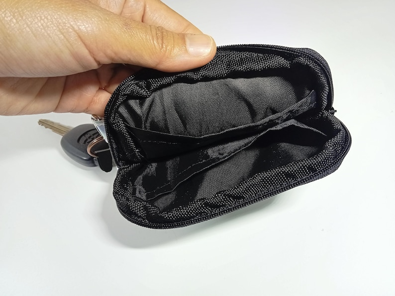 Peut inclure: Petite pochette &agrave; cl&eacute;s noire zipp&eacute;e ouverte, avec des poches int&eacute;rieures. Un jeu de cl&eacute;s est visible &agrave; c&ocirc;t&eacute; de la pochette ouverte. La pochette est en tissu textur&eacute; et r&eacute;sistant.