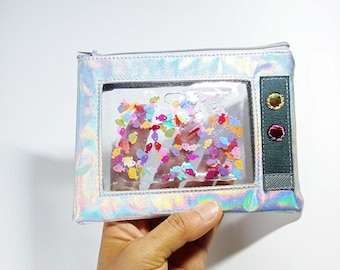 Peut inclure: Pochette rectangulaire irisée avec une fenêtre transparente remplie de confettis colorés en forme de poisson. La pochette a une fermeture éclair argentée et un panneau latéral gris foncé avec trois boutons décoratifs. La pochette est tenue dans une main.