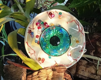 Peut inclure: Un sac cosmétique rond et transparent avec un grand motif d'œil détaillé. L'œil présente un iris vert, une pupille noire et des accents de paillettes bleues et vertes. Des détails rouges en forme de veines et des paillettes colorées décorent la surface du sac.