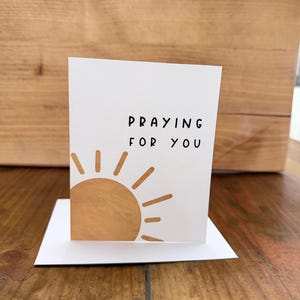 May include: A white greeting card with the text "PRAYING FOR YOU" in black. A tan sun graphic is on the bottom left. The card is standing on a white base, set against a wooden background.