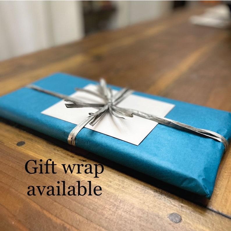 Pu&ograve; includere: Una scatola regalo blu turchese con un nastro argentato legato a fiocco. La scatola &egrave; avvolta in carta velina e ha un biglietto bianco in cima. Il testo "Gift wrap available" &egrave; scritto sotto la scatola.