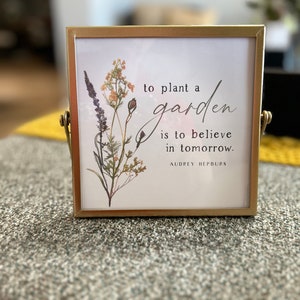 May include: A gold-framed picture with a white background and a floral illustration. The quote "To plant a garden is to believe in tomorrow." by Audrey Hepburn is printed on the picture.