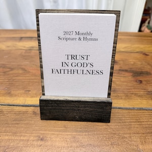 May include: A white rectangular card with black text reading 2027 Monthly Scripture & Hymns and TRUST IN GOD'S FAITHFULNESS, held upright by a dark brown stained wooden stand with a horizontal slot for the card to rest in.