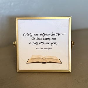 May include: A framed print with a gold-colored frame. The print features the quote "Nobody ever outgrows Scripture: the book widens and deepens with our years." by Charles Spurgeon, along with an illustration of an open book.