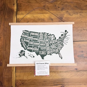 May include: A US Travel Map print with a wooden frame, displaying a dark green outline of the United States with state names. Includes a small packet with the text "US Travel Map" and instructions. The map is hanging on a wooden wall.