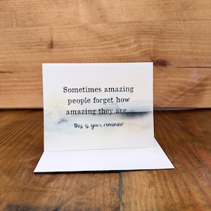 May include: A white greeting card with the text "Sometimes amazing people forget how amazing they are. This is your reminder." The card has a watercolor design in shades of blue and gray. The card is displayed on a wooden surface.