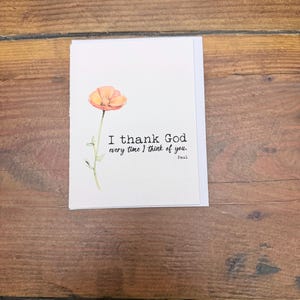 May include: A white greeting card featuring a watercolor illustration of a single orange poppy flower with a green stem and leaves. The text printed in black ink reads, I thank God every time I think of you. Paul.
