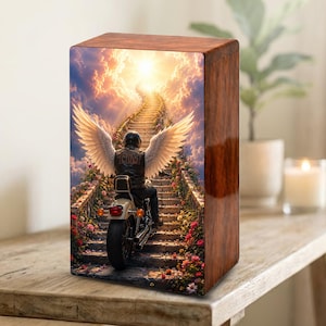 Può includere: Urna crematoria in legno con finitura lucida. La parte anteriore presenta l'immagine di una persona su una moto con ali d'angelo, che sale una scala verso una luce brillante nel cielo. L'urna è posta su una superficie di legno.