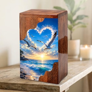 Könnte beinhalten: Holzurne mit glänzender Oberfläche. Die Vorderseite zeigt ein Bild von Möwen, die über einem Strand bei Sonnenuntergang fliegen, mit einer herzförmigen Wolkenformation. Die Urne steht auf einer Holzoberfläche.