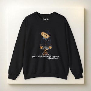 May include: Black crewneck sweatshirt featuring a graphic of a bear wearing a blazer, skirt, and boots. The text "POLO BEAR by RALPH LAUREN" is printed below the bear, with the designer's signature. The sweatshirt is a classic style.
