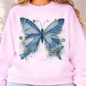 K&ouml;nnte beinhalten: Ein hellrosa Sweatshirt mit Rundhalsausschnitt und einem gro&szlig;en Schmetterlingsmotiv, das aus ausgefransten blauen Jeansstoff-Flicken besteht. Das Design enth&auml;lt kleine runde Kn&ouml;pfe, Ziern&auml;hte und eine blaue Stoffrose am unteren rechten Fl&uuml;gel.
