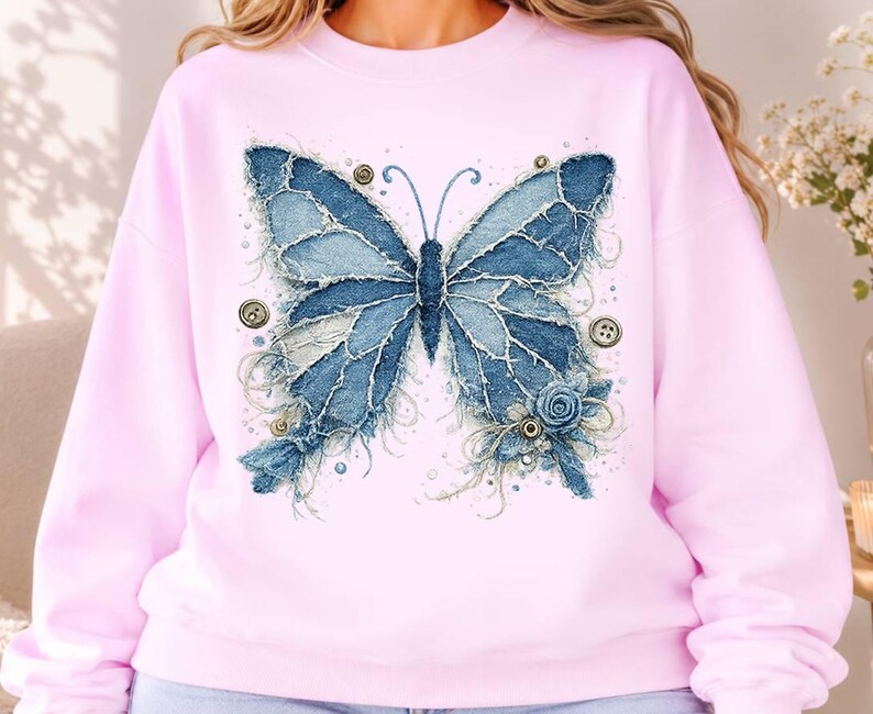 K&ouml;nnte beinhalten: Ein hellrosa Sweatshirt mit Rundhalsausschnitt und einem gro&szlig;en Schmetterlingsmotiv, das aus ausgefransten blauen Jeansstoff-Flicken besteht. Das Design enth&auml;lt kleine runde Kn&ouml;pfe, Ziern&auml;hte und eine blaue Stoffrose am unteren rechten Fl&uuml;gel.