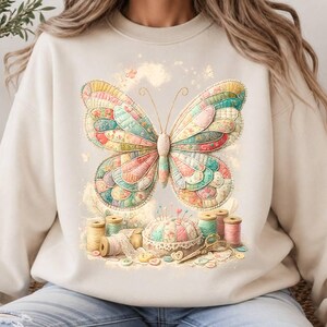 May include: A beige crewneck sweatshirt featuring a large, colourful butterfly graphic composed of quilted fabric patterns. Below the butterfly are sewing supplies including spools of thread, a round pincushion, lace trim, and small buttons.