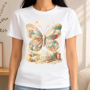 May include: A white short-sleeve t-shirt featuring a central graphic of a large, multi-coloured butterfly with a quilted texture. Below the butterfly are sewing supplies including several spools of thread, a round pincushion, and scissors.