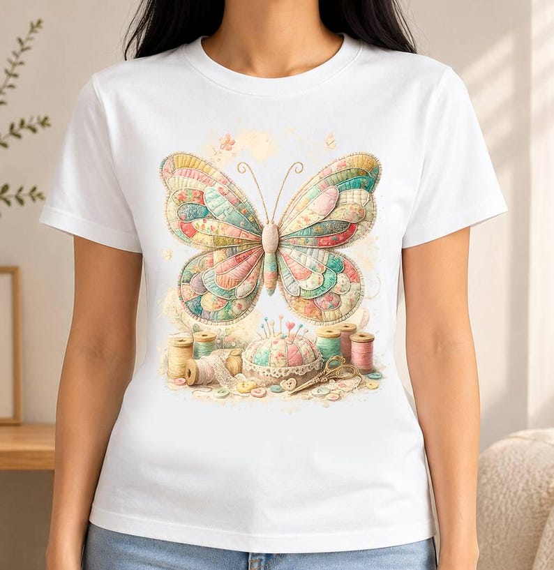 May include: A white short-sleeve t-shirt featuring a central graphic of a large, multi-coloured butterfly with a quilted texture. Below the butterfly are sewing supplies including several spools of thread, a round pincushion, and scissors.