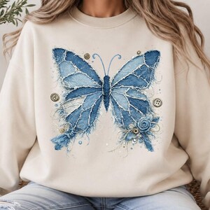 K&ouml;nnte beinhalten: Ein beigefarbenes Sweatshirt mit Rundhalsausschnitt und einer gro&szlig;en Grafik eines Schmetterlings, der aus ausgefransten blauen Jeansstoffst&uuml;cken zusammengesetzt ist. Das Design enth&auml;lt kleine runde Kn&ouml;pfe, eine Stoffrose und feine N&auml;hte.