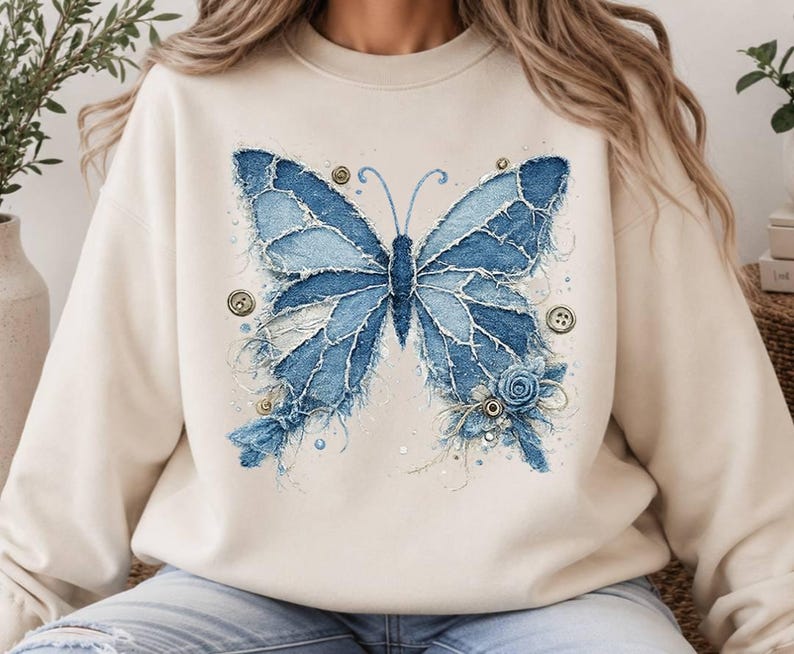 K&ouml;nnte beinhalten: Ein beigefarbenes Sweatshirt mit Rundhalsausschnitt und einer gro&szlig;en Grafik eines Schmetterlings, der aus ausgefransten blauen Jeansstoffst&uuml;cken zusammengesetzt ist. Das Design enth&auml;lt kleine runde Kn&ouml;pfe, eine Stoffrose und feine N&auml;hte.