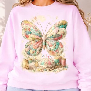 May include: A light pink crewneck sweatshirt featuring a large, colourful butterfly with a patchwork-style wing pattern. Below the butterfly, a sewing-themed graphic displays a pincushion, various spools of thread, lace trim, and scissors.