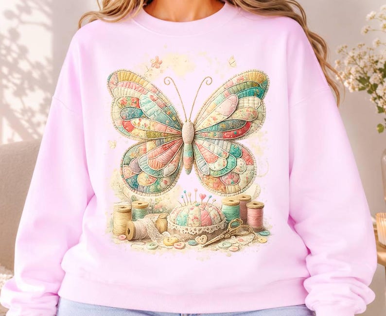 May include: A light pink crewneck sweatshirt featuring a large, colourful butterfly with a patchwork-style wing pattern. Below the butterfly, a sewing-themed graphic displays a pincushion, various spools of thread, lace trim, and scissors.