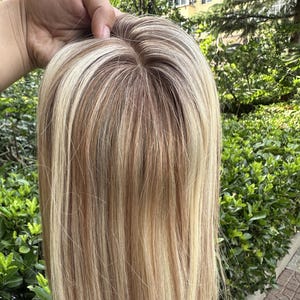 May include: A straight hair topper featuring a multi-tonal color palette with a blend of platinum blonde, light ash blonde, and warm honey brown highlights. The piece includes a visible monofilament base at the crown with a natural part.