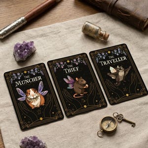 May include: Three rectangular tarot-style cards with black backgrounds and gold borders. The cards feature illustrations of a guinea pig labeled The Muncher, a mouse labeled The Thief, and a sugar glider labeled The Traveller, all with fairy wings.