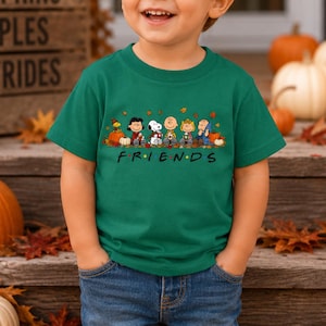 May include: A dark green short-sleeved t-shirt featuring a graphic print of the Peanuts characters sitting together with pumpkins and autumn leaves. Below the characters, the word FRIENDS is printed in black with colorful dots between letters.