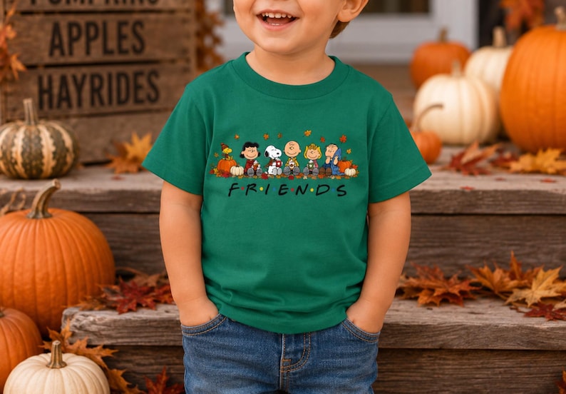 May include: A dark green short-sleeved t-shirt featuring a graphic print of the Peanuts characters sitting together with pumpkins and autumn leaves. Below the characters, the word FRIENDS is printed in black with colorful dots between letters.