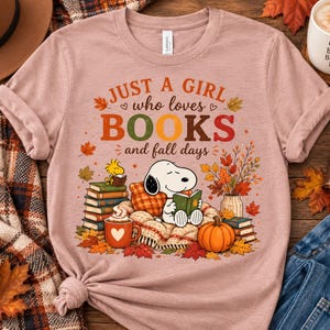 May include: A mauve short-sleeve t-shirt featuring a graphic of Snoopy and Woodstock reading books. The text reads, "JUST A GIRL who loves BOOKS and fall days." The design includes stacks of books, a mug, a pumpkin, and autumn leaves in orange and brown.