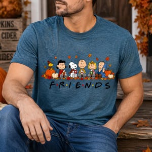 May include: A heathered blue short-sleeve t-shirt featuring a graphic of the Peanuts characters sitting together among autumn leaves and pumpkins. Below the characters, the word FRIENDS is printed in black with a red maple leaf as the letter I.