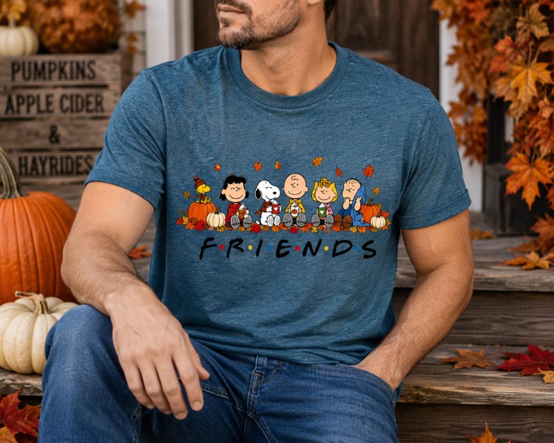 May include: A heathered blue short-sleeve t-shirt featuring a graphic of the Peanuts characters sitting together among autumn leaves and pumpkins. Below the characters, the word FRIENDS is printed in black with a red maple leaf as the letter I.