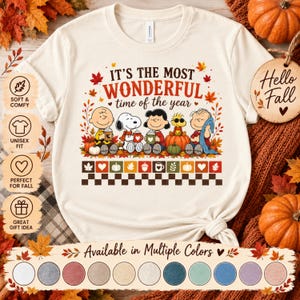 May include: A cream-colored t-shirt featuring a graphic of Peanuts characters with pumpkins and autumn leaves. The text reads, "IT'S THE MOST WONDERFUL time of the year." Below the characters is a row of icons including leaves, hearts, and mugs, followed by a checkered pattern.