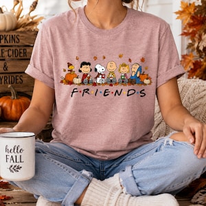May include: A mauve short-sleeve t-shirt featuring a graphic of the Peanuts characters sitting among autumn leaves and pumpkins. Below the characters, the word FRIENDS is printed in black with colorful dots between the letters. A white mug with the text hello FALL is held.