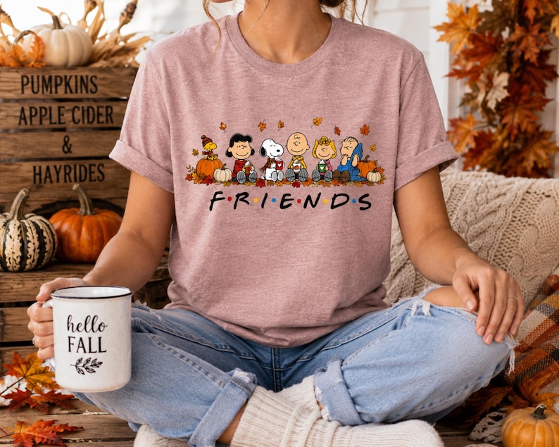 May include: A mauve short-sleeve t-shirt featuring a graphic of the Peanuts characters sitting among autumn leaves and pumpkins. Below the characters, the word FRIENDS is printed in black with colorful dots between the letters. A white mug with the text hello FALL is held.