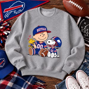 May include: A heather gray crewneck sweatshirt featuring a graphic print of Charlie Brown, Snoopy, and Woodstock wearing blue and red Buffalo football jerseys and helmets. Charlie Brown wears a blue cap with the word BUFFALO printed in white.