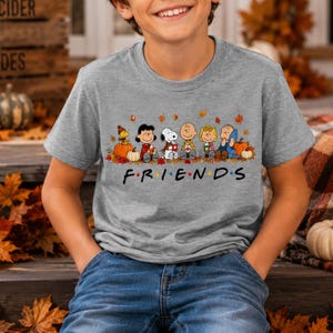 May include: A heather gray short-sleeve t-shirt featuring a graphic of Peanuts characters sitting among autumn leaves and pumpkins. Below the characters, the text FRIENDS is printed in a black font with colorful dots between each letter.