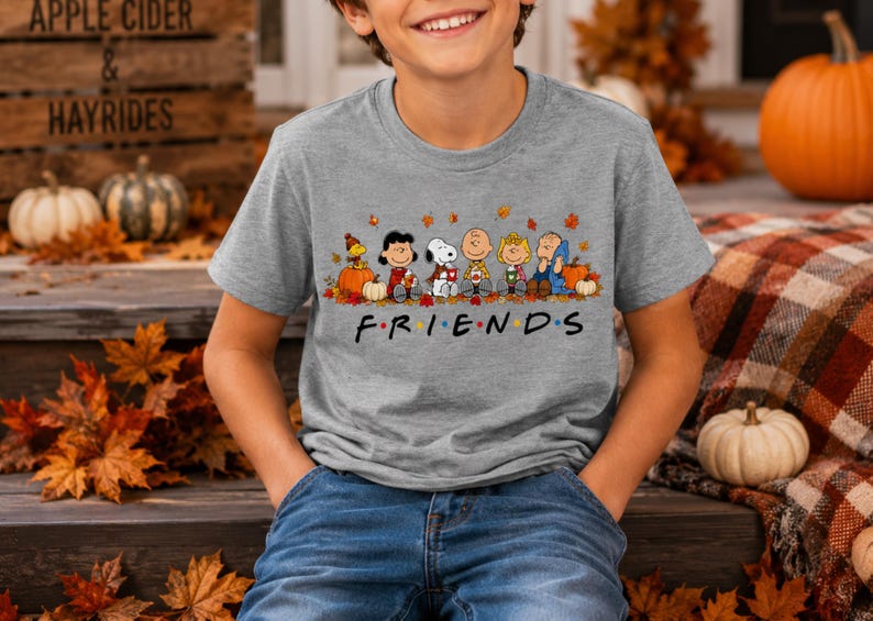 May include: A heather gray short-sleeve t-shirt featuring a graphic of Peanuts characters sitting among autumn leaves and pumpkins. Below the characters, the text FRIENDS is printed in a black font with colorful dots between each letter.