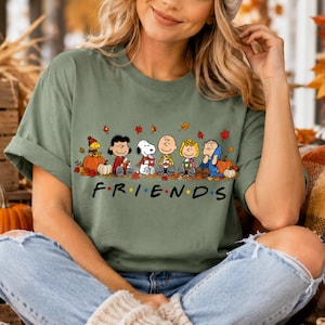 May include: An olive green short-sleeve t-shirt featuring a graphic print of the Peanuts characters sitting together among autumn leaves and pumpkins. Below the characters, the word FRIENDS is printed in black with colorful dots.