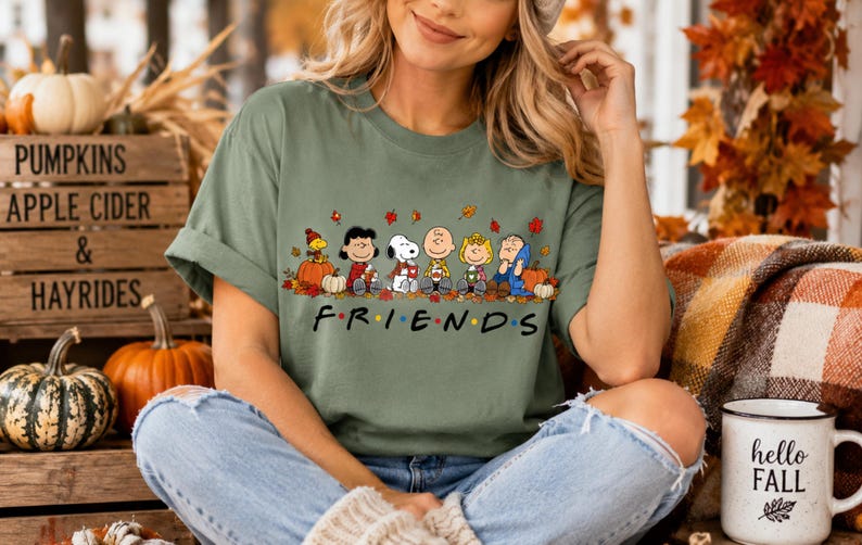 May include: An olive green short-sleeve t-shirt featuring a graphic print of the Peanuts characters sitting together among autumn leaves and pumpkins. Below the characters, the word FRIENDS is printed in black with colorful dots.