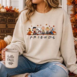 May include: A beige crewneck sweatshirt featuring a graphic of the Peanuts characters sitting together among autumn leaves and pumpkins. Below the characters, the word FRIENDS is printed in black with colorful dots between the letters.
