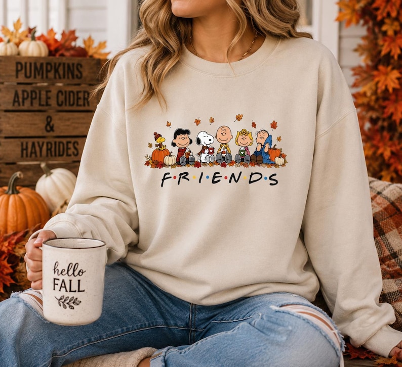 May include: A beige crewneck sweatshirt featuring a graphic of the Peanuts characters sitting together among autumn leaves and pumpkins. Below the characters, the word FRIENDS is printed in black with colorful dots between the letters.