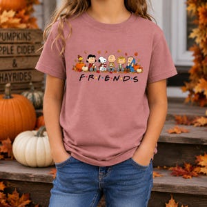 May include: A mauve short-sleeve t-shirt featuring a graphic of Peanuts characters sitting among pumpkins and autumn leaves. Below the characters, the text reads "F R I E N D S" with colorful dots between the letters, styled like a television show logo.