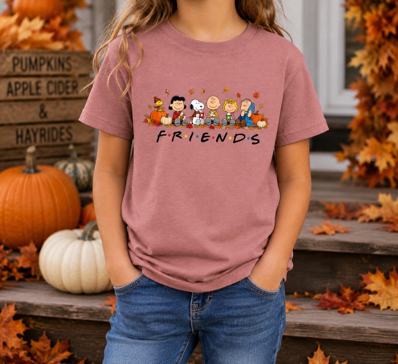 May include: A mauve short-sleeve t-shirt featuring a graphic of Peanuts characters sitting among pumpkins and autumn leaves. Below the characters, the text reads "F R I E N D S" with colorful dots between the letters, styled like a television show logo.