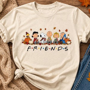 May include: A beige short-sleeve t-shirt featuring a graphic of the Peanuts characters sitting together among autumn leaves and pumpkins. Below the characters, the word FRIENDS is printed in black with colorful dots between each letter.