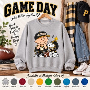 May include: A light gray crewneck sweatshirt featuring a graphic of Charlie Brown and Snoopy wearing Pittsburgh football gear. The text reads GAME DAY Looks Better Together, Good Friends Football & Happy Days. The design includes a football.
