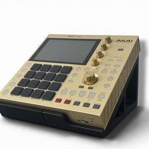 Puede incluir: Un controlador de producción musical Akai Professional MPC One dorado con pantalla táctil negra, dieciséis pads cuadrados negros, varios botones plateados y cuatro perillas giratorias doradas. El dispositivo está sobre un soporte negro.