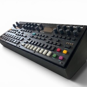 Op de afbeelding: Een zwarte Elektron Digitone synthesizer met een donker bedieningspaneel met talloze knoppen, een centraal display en een raster van toetsen, waaronder witte, rode, gele, groene en paarse knoppen voor geluidsmanipulatie en sequencing.