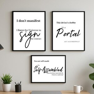 May include: Three framed art prints with black frames and white backgrounds. The prints feature black text with phrases like "I don't manifest" and "This lab isn't a hobby." The prints are displayed above a wooden desk with a laptop and a small plant.