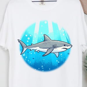 May include: White t-shirt with a cartoon shark design. The shark is grey and white, set against a blue underwater scene. The shirt is hanging on a wooden hanger.