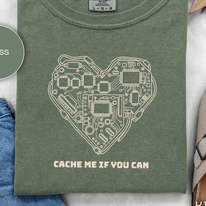 May include: A moss green t-shirt featuring a heart-shaped circuit board design and the text "CACHE ME IF YOU CAN". The shirt is a Comfort Colors brand, size L-G-G.