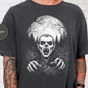 Könnte beinhalten: Pfefferfarbenes T-Shirt mit einer schwarz-weißen Grafik eines Vampirs. Das Design zeigt einen Vampir mit offenem Mund, ausgestreckten Händen und einem Vollmondhintergrund mit Rosen und Bäumen. Der Text "KITTED KOOL" befindet sich unten rechts.
